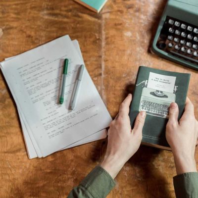 A vintage setup showcasing a typewriter, manuscripts, and hands holding a book on writing.
